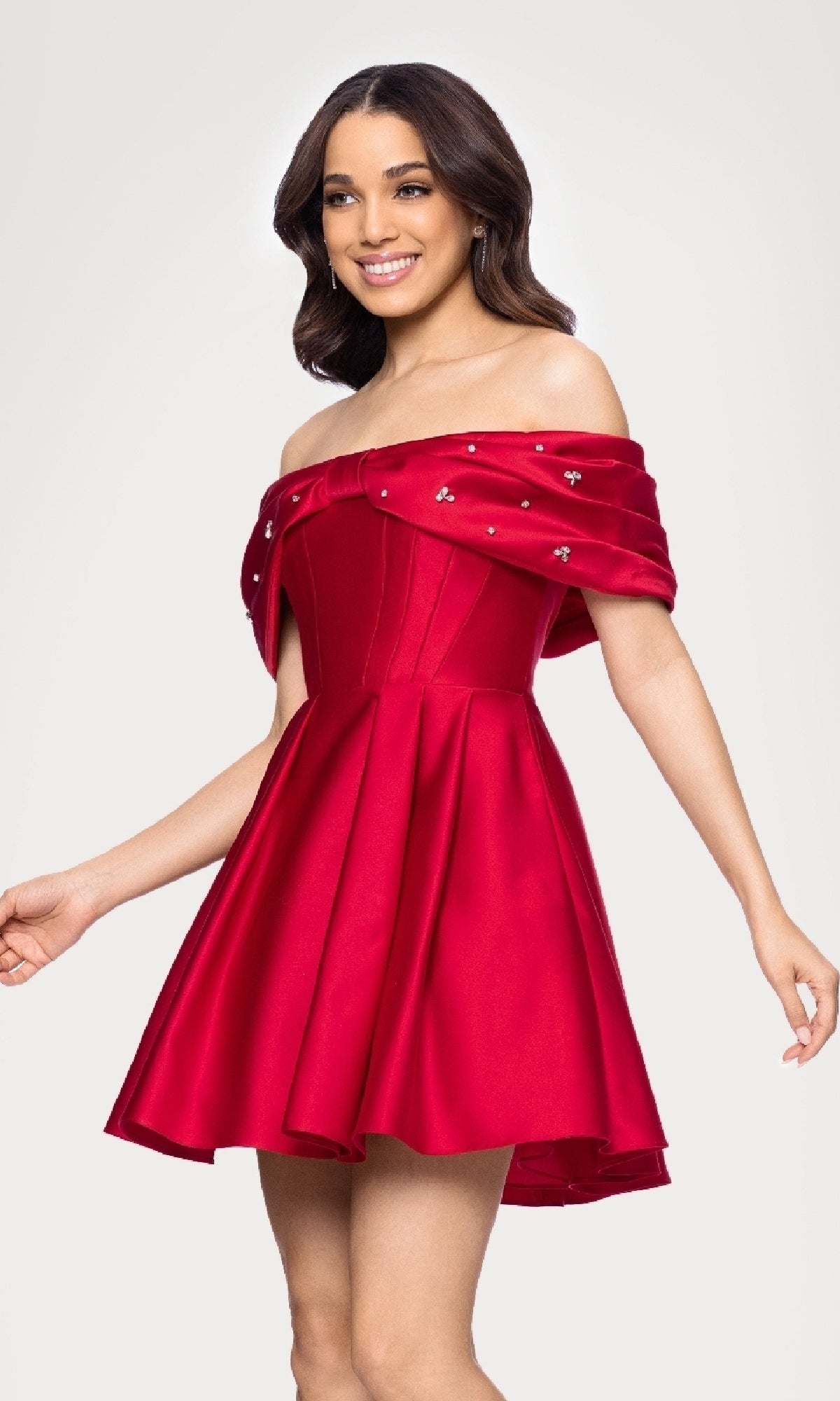 A woman smiles in the Off-Shoulder Red Hoco Dress: Blondie Nites 5695BN, featuring a short red skirt, corset-style bodice, and embellished off-the-shoulder neckline, standing against a plain background.
